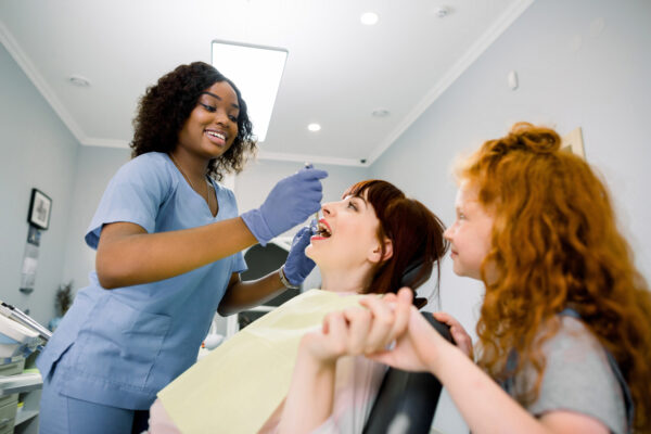 Side bottom view of young Caucasian woman having teeth checkup and treatment. Her little daughter holds hand of mom, while female black dentist with tools provides teeth cure.