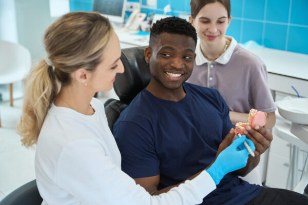 Smiling African American client satisfied with dental prosthetics way the stomatologist and nurse advising to him, aesthetic dentistry