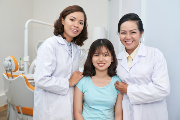 Cheerful Asian dentists and charming girl in modern dental office smiling happily at camera